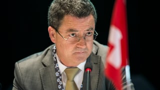 Rossier sitzt vor einem Mikrofon, unscharf im Bild auch eine kleine Schweizerfahne.