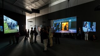 Menschen stehen vor Filmleinwänden in einem Ausstellungsraum.