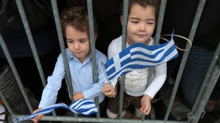 Zwei Kinder schwenken hinter einer Absperrung griechische Fahnen.