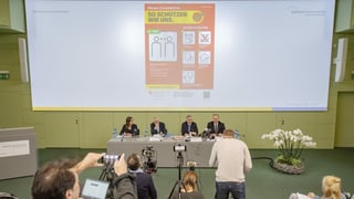Vier Personen sitzen vor einer Leinwand mit Warnhinweisen. Vier Personen sitzen vor einer Leinwand mit Warnhinweisen.