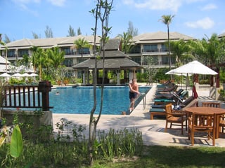 Eine Hotelanlage, eine Person steigt aus dem Wasser, dahinter sind merstöckige Bungalos zu sehen.
