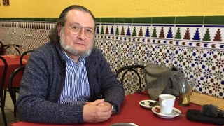 Der Ökonom Santiago Niño Becerra sitzt an einem Tisch. Er hat einen Bart und trägt eine Brille. Vor ihm auf dem Tisch steht eine weisse Kaffeetasse. Der Hintergrund ist recht farbig, das Tischtuch rot. 