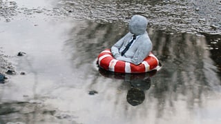 Die Skulptur eines kleinen Männchens in einem Schwimmring dümpelt in einer Pfütze.
