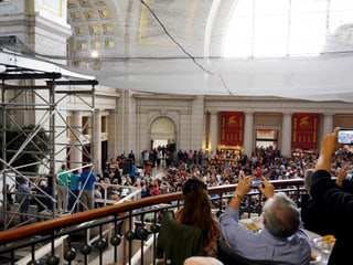 In einer Halle sind viele Menschen versammelt, die auf eine kleine Bühne schauen. In einer Halle sind viele Menschen versammelt, die auf eine kleine Bühne schauen.