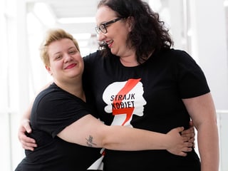 Zwei jüngere Frauen in schwarzen T-Shirts umarmen sich und lachen.