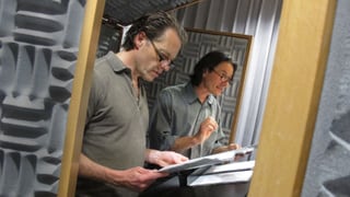 Blick durch die Tür ins Hörspielstudio auf Oliver Sauer und Till Kretzschmar mit Manuskript in der Hand vor dem Mikrofon.