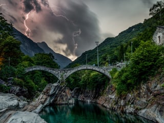 Steiniger Fluss mit alter Steinbrücke. Im Hintergrund dunkle Wolken mit Blitzen. Es scheint Dämmerung zu sein. 
