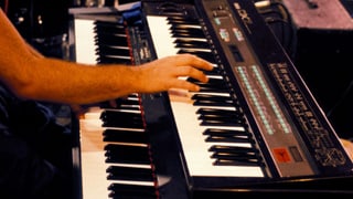 Eine Hand greift in die Tasten eines Synthesizers mit dem Label DX7. Eine Hand greift in die Tasten eines Synthesizers mit dem Label DX7.