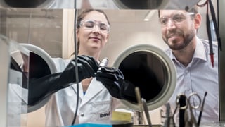 Zwei Wissenschaftler vor einem Glaskasten, die Frau hält mit Handschuhen ein Röhrchen.