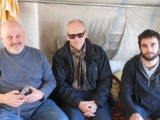 Zu sehen sind drei Männer, wie sie in einem Zelt sitzen. Gekleidet sind sie leger in Pullover und Jeans. Der Mann ganz links trägt einen kurzen weissen Bart und Glatze, der Mann in der Mitte trägt Sonnenbrille, der Mann rechts dunkle kurze Haare und Bart.