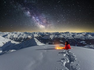 Nachtbild in den sternenklaren Alpen mit Schnee und gut sichtbarer Milchstrasse. Im Schnee sitzender Mensch.