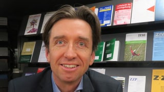 Ein Mann mit braunen Haaren vor einem Büchergestell.