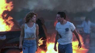 Eine Frau (links) und ein Mann (rechts) laufen in zerfetzten Kleidern von einem brennenden Auto weg.