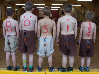 Fünf Jungs stehen mit nacktem Oberkörper in einem Fensterrahmen und haben Buchstaben auf den Rücken gemalt. 