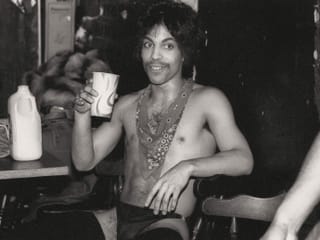 Ein Foto von Prince von 1985: Er sitzt halbnackt in einem Stuhl und trinkt aus einem Becher.