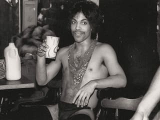 Ein Foto von Prince von 1985: Er sitzt halbnackt in einem Stuhl und trinkt aus einem Becher. Ein Foto von Prince von 1985: Er sitzt halbnackt in einem Stuhl und trinkt aus einem Becher.