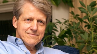 Portrait von Robert Shiller auf einem blauen Sessel, im Hintergrund Blätter einer Pflanze. Portrait von Robert Shiller auf einem blauen Sessel, im Hintergrund Blätter einer Pflanze.