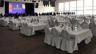 Ein Saal mit weiss gedeckten Tischen, im Hintergrund Menschen vor einer Leinwand Ein Saal mit weiss gedeckten Tischen, im Hintergrund Menschen vor einer Leinwand