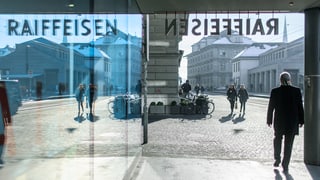Raiffeisen-Schriftzug an einem Gebäude spiegel sich in einer Glasfrobt, ein Mann geht vorbei.