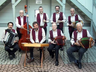 Acht Musikanten mit ihren Instrumenten auf dem Gruppenfoto vor einem weissen Holzhaus.