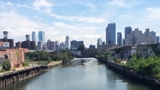 Ein Kanal fliesst duch ein Industriegebiet. Im Hintergrund die Skyline von New York. 