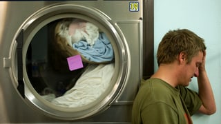 Ein junger Mann lehnt sich an eine öffentliche Waschmaschine und hält sich die Hand an die Stirn.