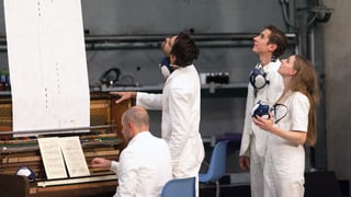 Vier Menschen in weissen Anzügen: Drei stehen und blicken in die Höhe, einer sitzt am Klavier.