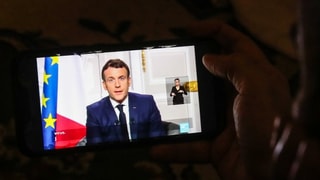 Hand hält Tablet. Darauf ist Macron zu sehen, der eine Ansprache hält.