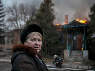 Eine Frau blickt in die Kamera, hinter ihr ein brennendes Haus.