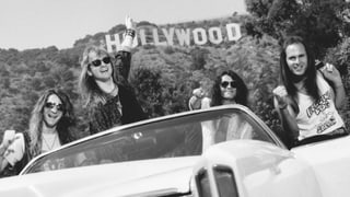 Schwarzweissfoto: Vier Männer mit langen Haaren und Sonnenbrillen posieren in einem Cabrio vor dem "Hollywood"-Schild.