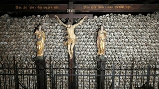 Unzählige, aufeinandergsetapelte Totenschädel. Davor Christuskreuz, darüber ein Holzbalken mit Inschrift.