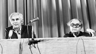 Die Schriststeller Adolf Muschg und Max Frisch auf einer Aufnahme von 1975 an einem Tisch mit Mikrofonen.