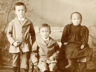 Zwei Jungen und ein Mädchen, alle Geschwister, schauen adrett gekleidet direkt in die Kamera.