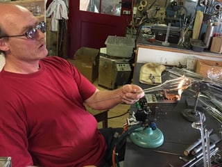 Ein Mann hält ein Stück Glas in eine Flamme und trägt eine Schutzbrille.