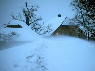 Zwei Bauernhäuser tief im Schnee. In einem Fenster leuchtet ein kleines Licht. 