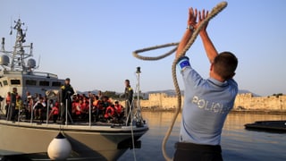 Ein Polizist wirft ein Seil zu einem Küstenwache-Boot voller Flüchtlinge.