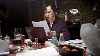 Sherlock liest an einem chaotischen Tisch handgeschriebene Dokumente.