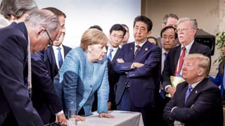 Trump sitzt mit verschränkten Armen, Merkel versucht ihn zu überzeugen