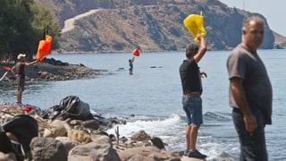 Menschen stehen am Strand und winken mit farbigem Plastik in den Händen aufs Meer hinaus. 
