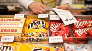 Auslage eines Kiosks mit Toblerone, M&Ms und Maltesers. Eine Frau prüft einen Kassenbon.