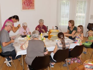 An einem Tisch voller Bastelmaterialien sitzen Menschen verschiedenen Alters.