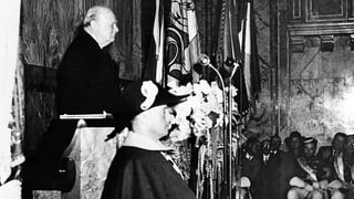 Ein älterer Herr steht an einem Rednerpult und blickt ins Publikum. Ein älterer Herr steht an einem Rednerpult und blickt ins Publikum.
