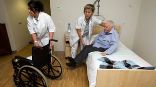Pflegerinnen in einem Pflegeheim helfen einem pflegebedürftigen Senior aus dem Bett.