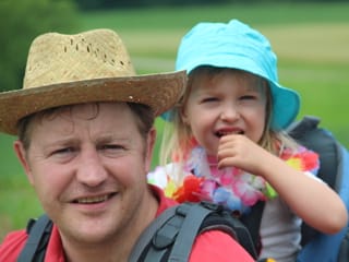 Vater und Tochter. Die Tochter wird im Rucksack auf Papas Rücken getragen und strahlt.