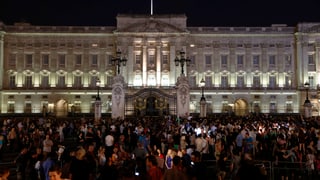 Beleuchteter Buckingham Palace am Abend, davor eine Menschenmasse.