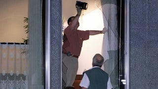Zwei Männer untersuchen ein eingeschlagenes Fenster mit einer Lampe.