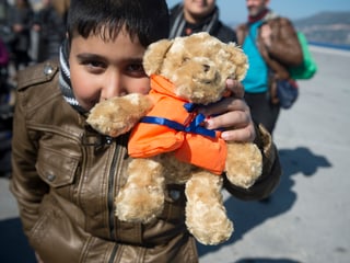 Ein Junge hält sich einen Teddybär mit orangefarbener Schwimmweste vor das Gesicht.