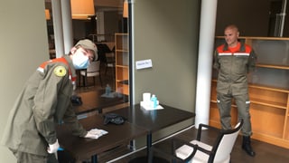 Ein junger Mann mit Mundschutz in der orangen Zivilschutzuniform putzt einen Tisch in einem kleinen Raum. Ein junger Mann mit Mundschutz in der orangen Zivilschutzuniform putzt einen Tisch in einem kleinen Raum.