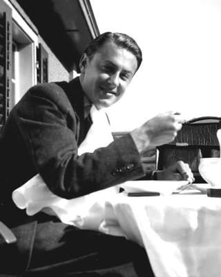 Artur Beul sitzt draussen vor einem Chalet an einem Tisch. Er ist hübsch frisiert und trinkt eine Tasse Kaffee.