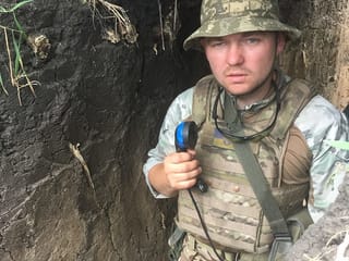 Ein junger Mann in Soldaten-Kluft steht in einer Art Schützengraben udn hält einen Telefonhörer in der Hand.